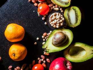 A vibrant arrangement of fruits and nuts on a black background featuring kiwi, avocado, oranges, apple, and peanuts.