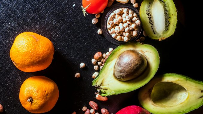 A vibrant arrangement of fruits and nuts on a black background featuring kiwi, avocado, oranges, apple, and peanuts.