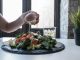 Hand pouring dressing over fresh salad with kale and tomatoes in a modern Dubai restaurant.