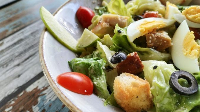 A close-up of a Caesar salad featuring fresh lettuce, eggs, croutons, and olives on a rustic table.