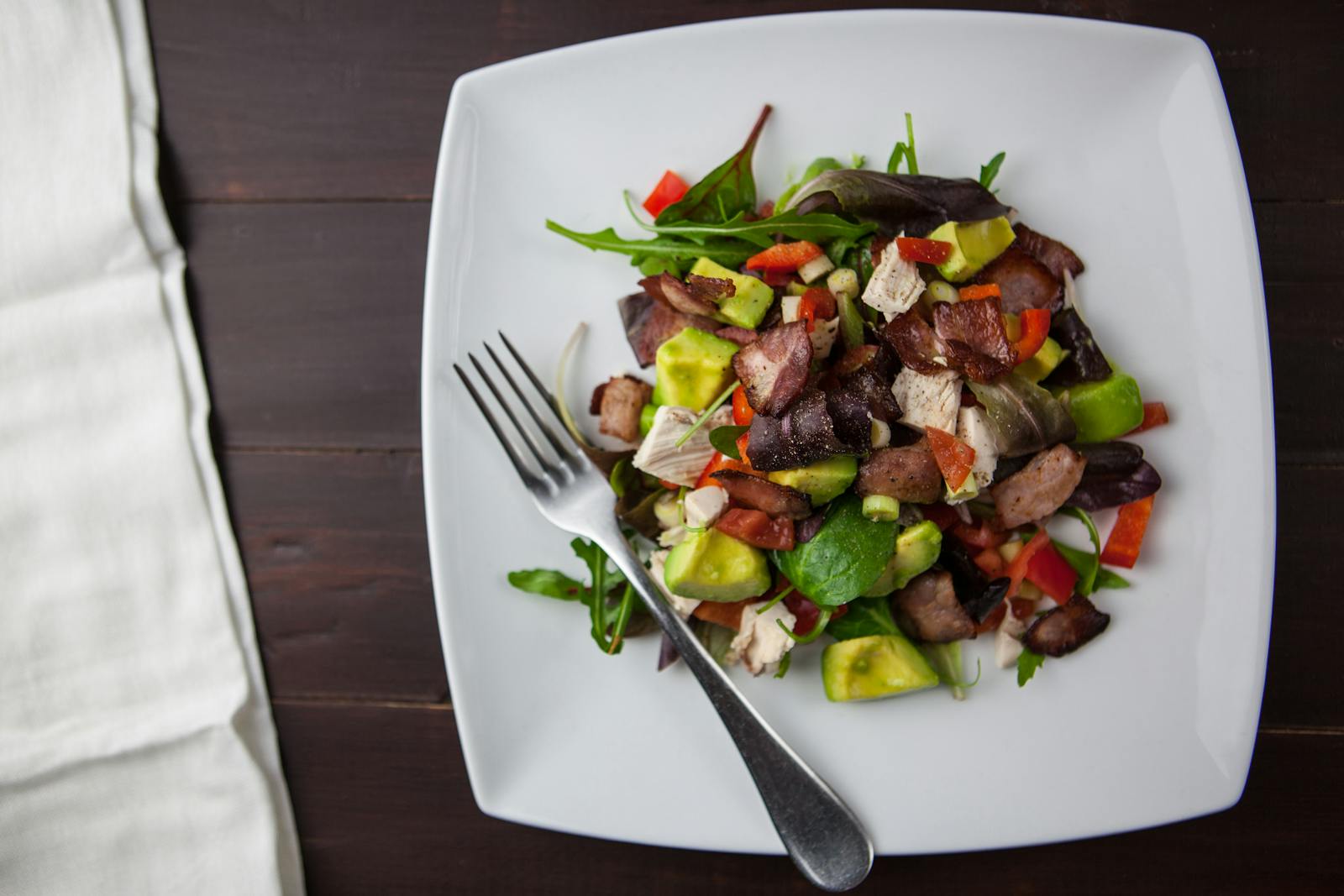 A fresh bacon, avocado, and mixed greens salad served on a square plate with a fork.